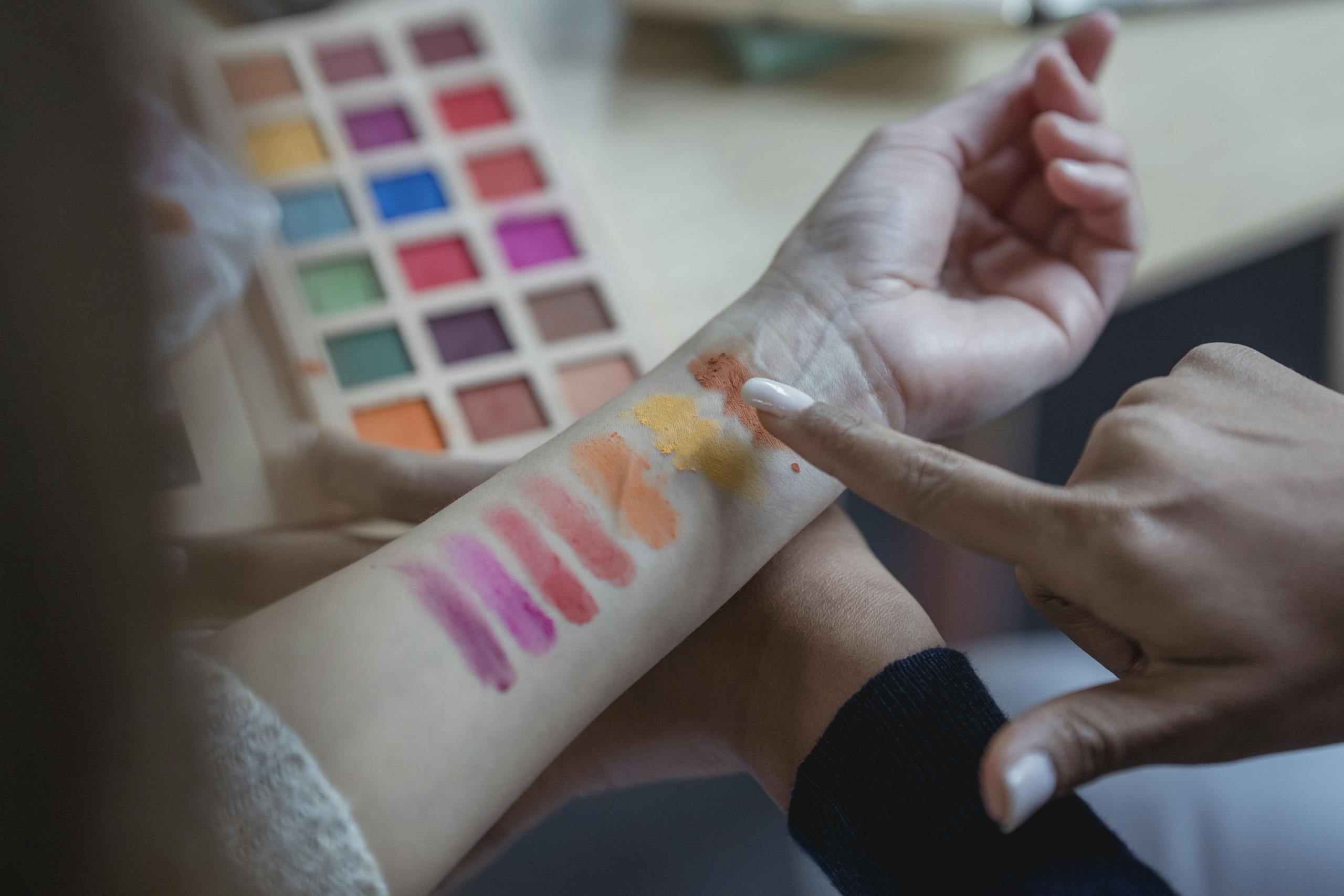 From above of crop unrecognizable young female makeup artist pointing at arm of client while applying colorful eyeshadows on wrist for testing